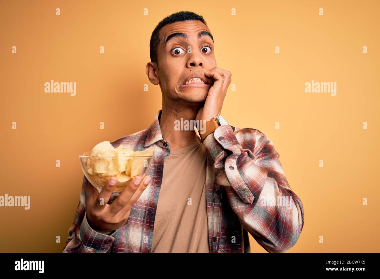 Junger gutaussehender afroamerikanischer Mann, der mit der Hand am Kopf eine Schüssel mit Kartoffelchips über gelbem Hintergrund hält, schockiert mit Scham und Überraschung Stockfoto