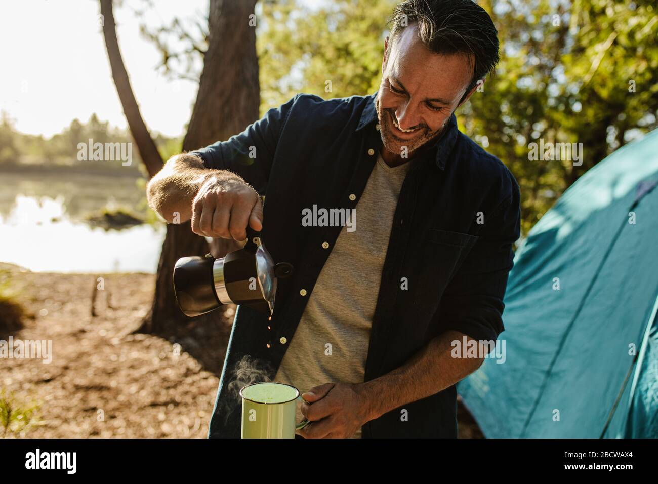 Lächelnder reifer Mann, der Kaffee aus dem Wasserkocher in eine Tasse gießt. Ein mittelgroßer Erwachsener, der auf dem Camping am See frischen Kaffee serviert. Stockfoto