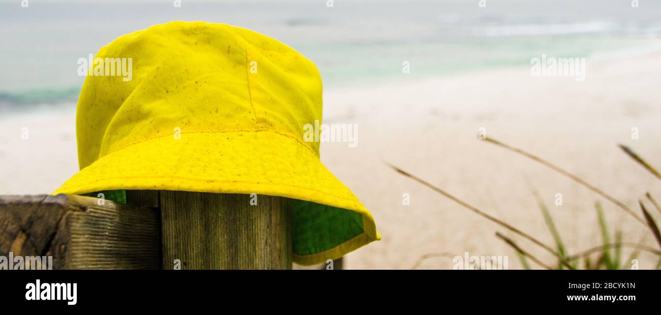 Gelber Sonnenhut aus Stoff ruht auf dem Pfosten am weißen Sandstrand mit sanften Wellen. Banner für Sommerferienkonzept, Flucht und Ungeweckte Kurzurlaube. Stockfoto