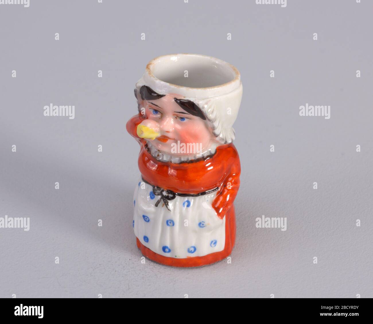 Matchsafe. Forschung in ProgressFigur der Frau, die aus der gelben Flasche trinkt. Figur trägt ein Rostkleid mit einer weißen Rüsche um den Hals und einer weißen Schürze mit großen blauen Punkten. Die Hand der Figur befindet sich in einer Schürzentasche. Matchsicher Stockfoto