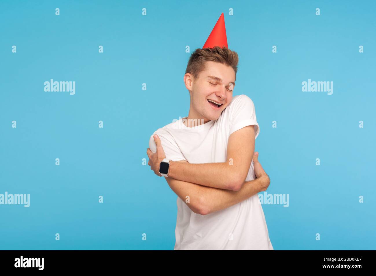 Alles gute zum Geburtstag! Lustiger positiver Mann mit Party-Kegel auf dem Kopf, der sich mit Freuden umarmt, gerne allein Urlaub feiert, sel Stockfoto