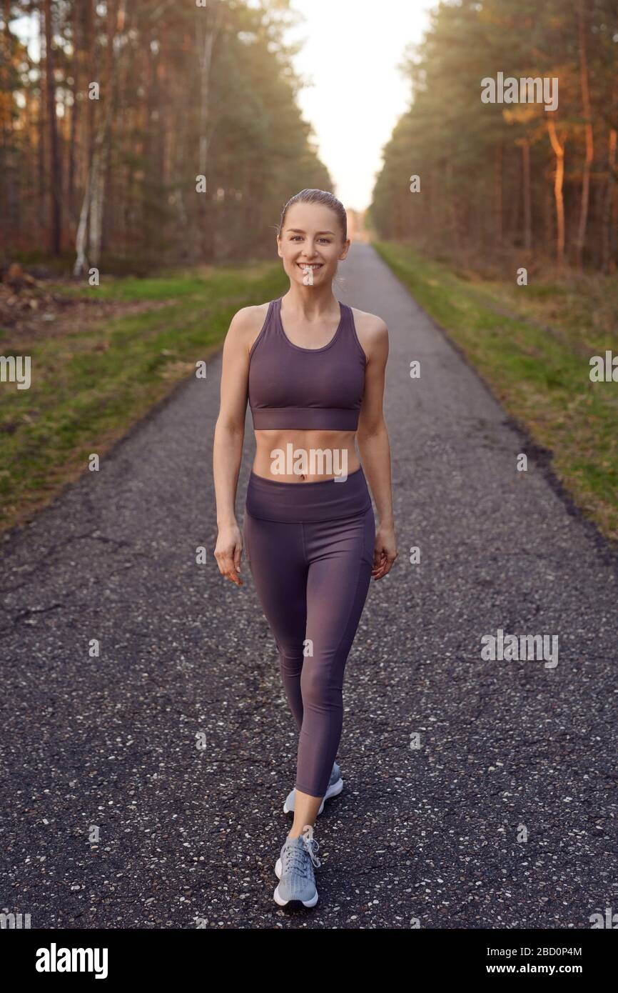 Lächelnde, sportliche Passform junge Frau, die auf einer geteppelten Spur durch Wälder arbeitet, die durch das warme Leuchten der Sonne in einem gesunden, aktiven Lifestyle-Schein beleuchtet werden Stockfoto