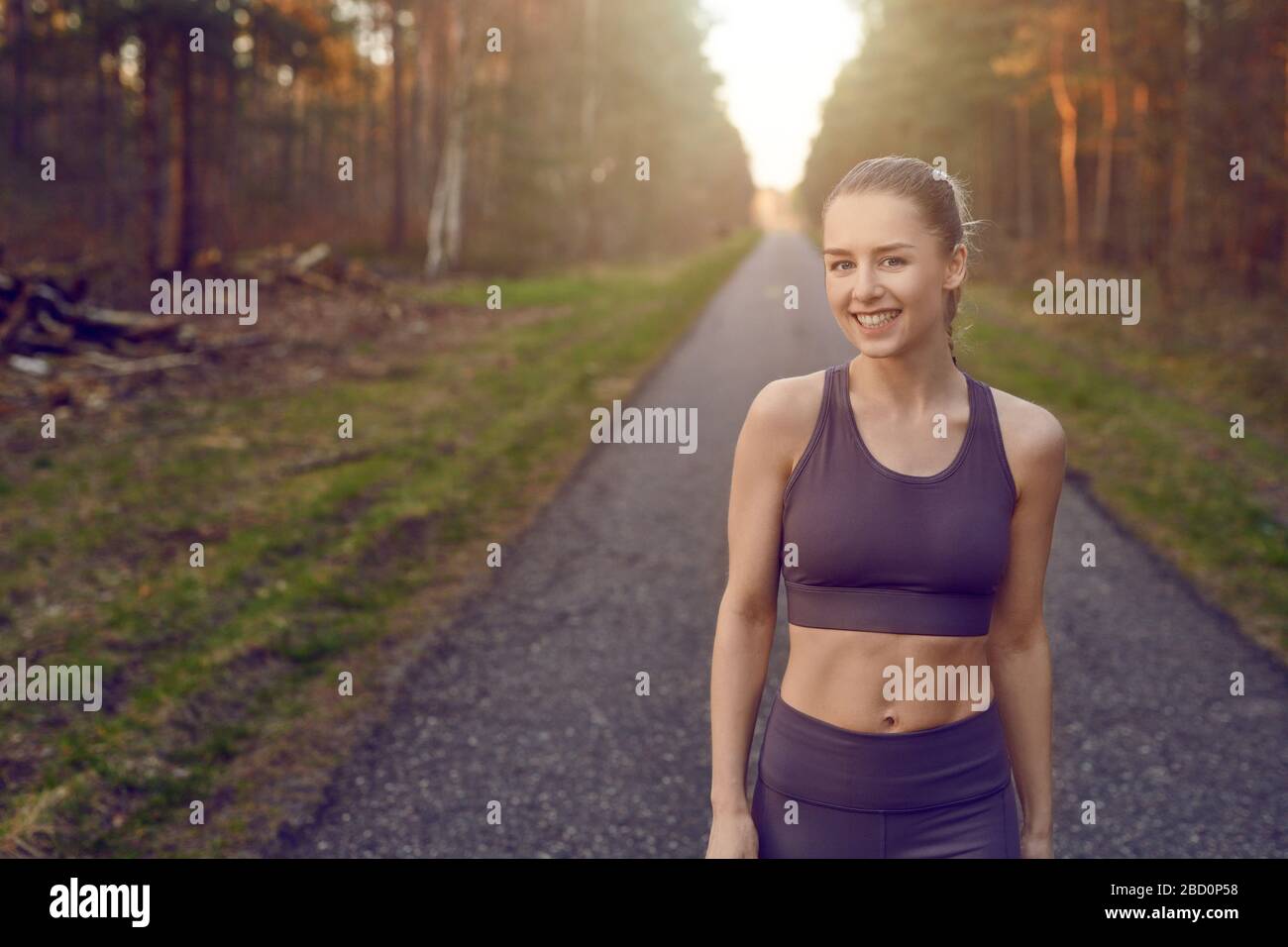 Lächelnde, sportliche Passform junge Frau, die auf einer geteppelten Spur durch Wälder arbeitet, die durch das warme Leuchten der Sonne in einem gesunden, aktiven Lifestyle-Schein beleuchtet werden Stockfoto