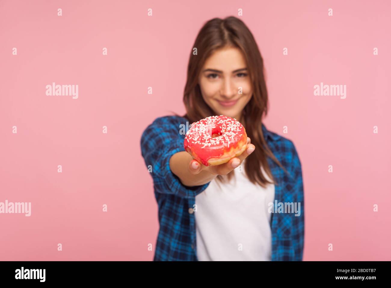 Portrait des positiven Mädchens im karierten Hemd, das leckeren Donut aushält und mit einem Lächeln auf die Kamera lächelt und gesüßte Süßwaren zeigt, die hoch in Glukose und Kalori sind Stockfoto