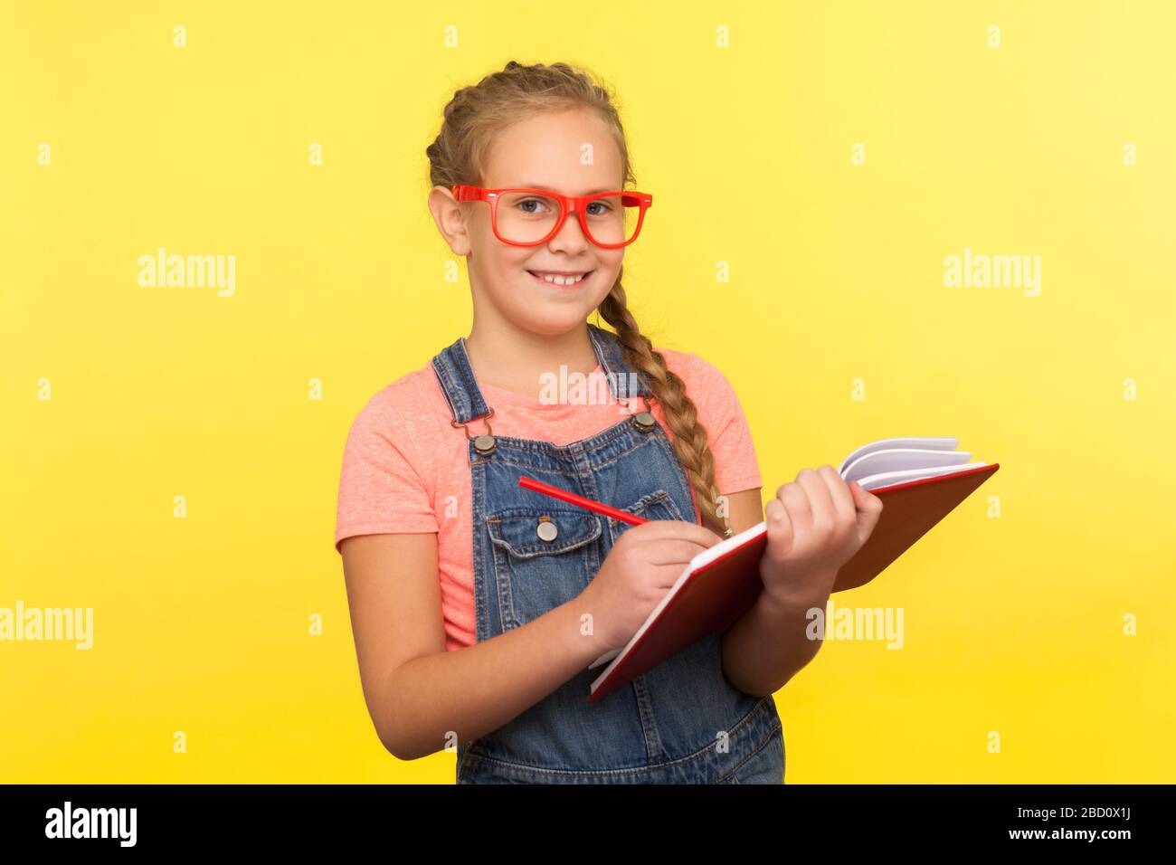 Porträt eines glücklichen, cleveren kleinen Mädchens in roter Brille, das Notizbuch und Bleistifte hält, mit der Kamera lächelt, kreative Ideen schreibt und interessante Lehrbücher genießt Stockfoto