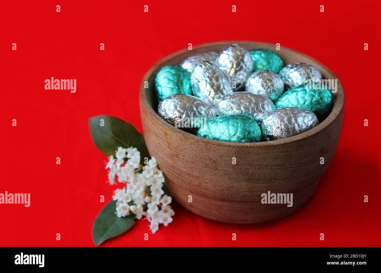 Eine mit Silber und Aqua-Grünfolie gefüllte wollige Schüssel bedeckte ostereier. In Nahaufnahme vor einem leuchtend orange roten Hintergrund mit weißer Frühlingsblume. Stockfoto