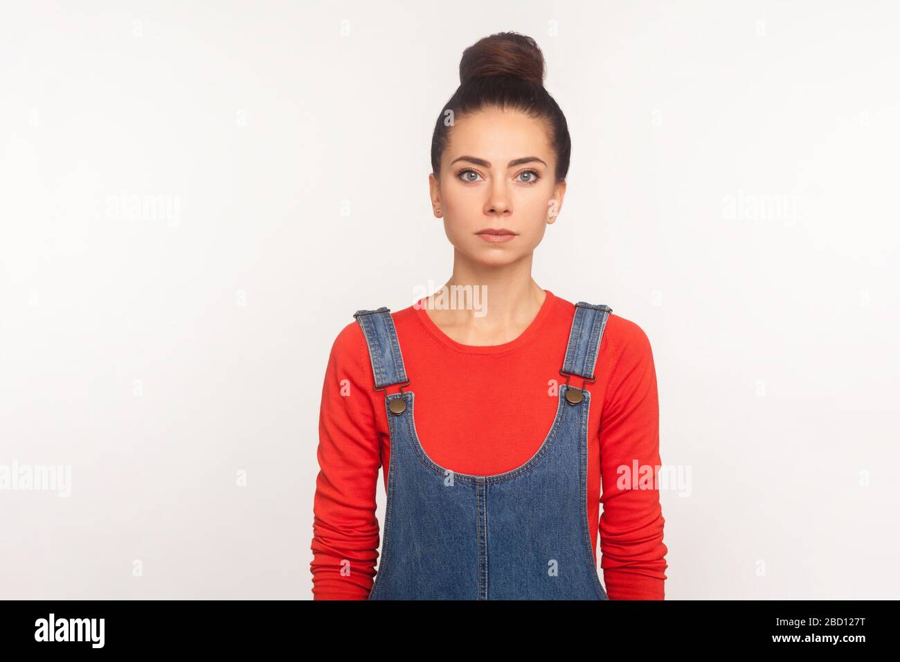 Portrait des eleganten, schlanken Mädchens mit Haarbüschen in Denim-Overalls stehend, Blick auf Kamera mit ernstem Gesicht, ruhiger, auffälliger Gesichtsausdruck. i Stockfoto