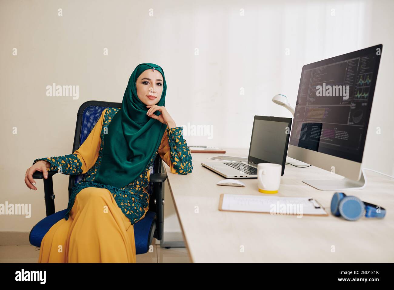 Selbstbewusste muslimische Softwareentwicklerin im traditionellen Kleid in Hijab, die an ihrem Bürotisch sitzt Stockfoto