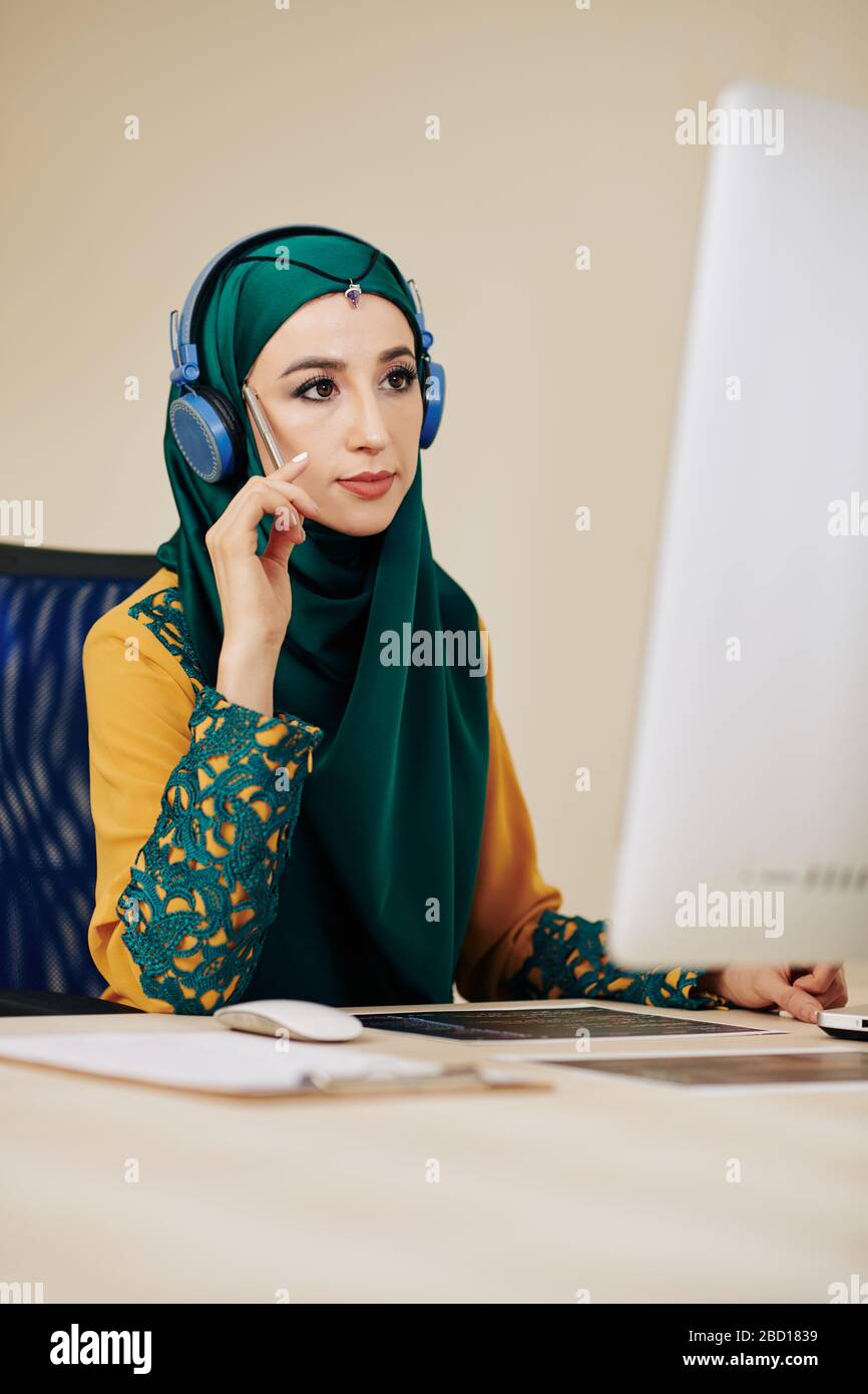 Pensive muslimische Programmiererin im traditionellen Kleid mit Blick auf den Bildschirm des Computers Stockfoto