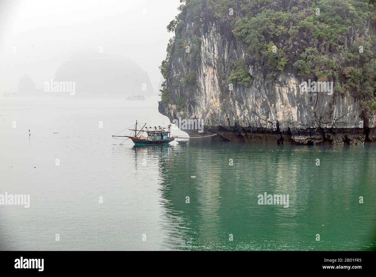Fischerboot neben einer Kalksteininsel mit grünem Meer an einem nebligen Tag in Halong Bay, Vietnam Stockfoto