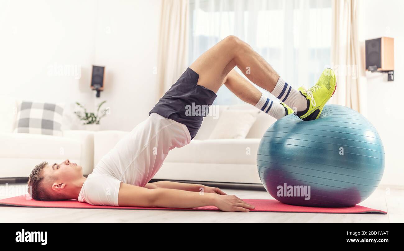 Junge Männer, die zu Hause auf einer roten Matte mit einem blauen Fit Ball trainieren und dabei Gluten, Beine und Körperkern trainieren. Stockfoto