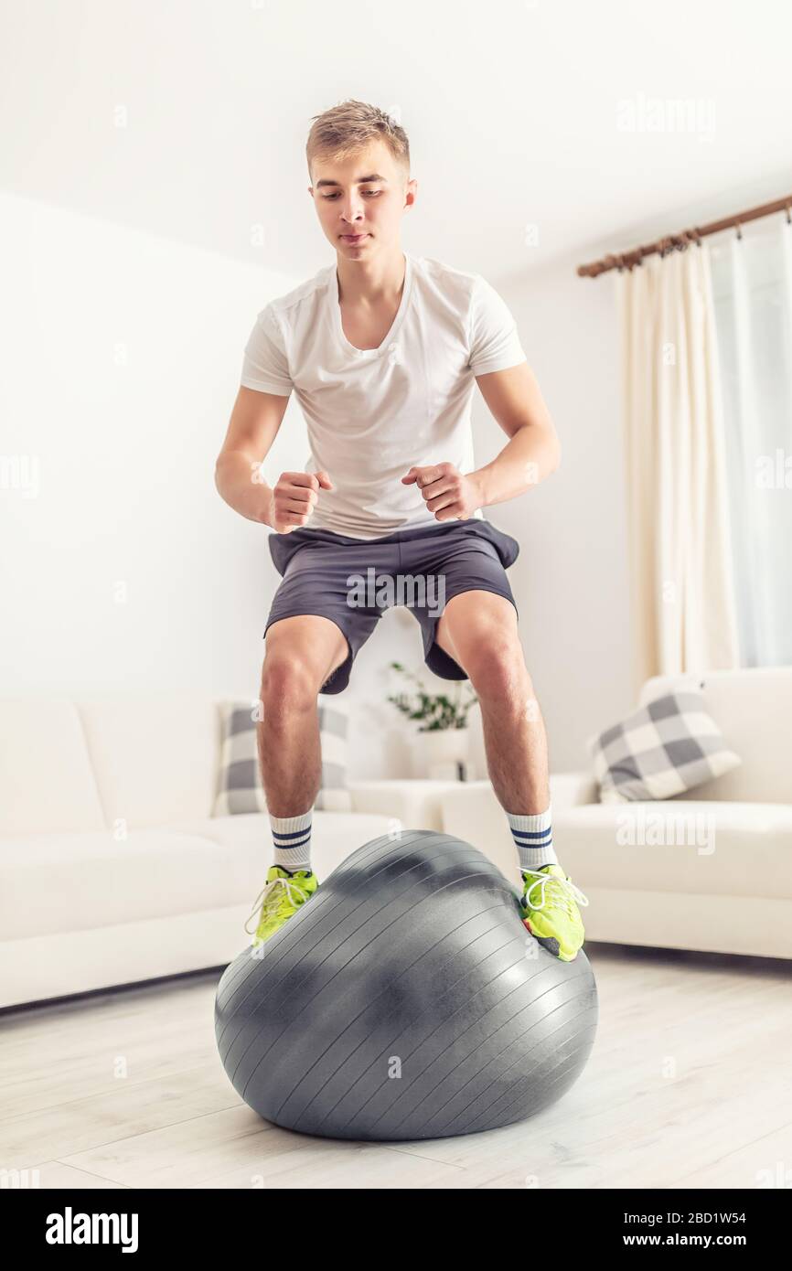 Konzentrierter junger Athlet, der während seines Heimtrainings im Schlafzimmer auf halbdeflationiertem grauem Fitball steht, der Fäuste geschlossen hält und nach unten blickt. Stockfoto