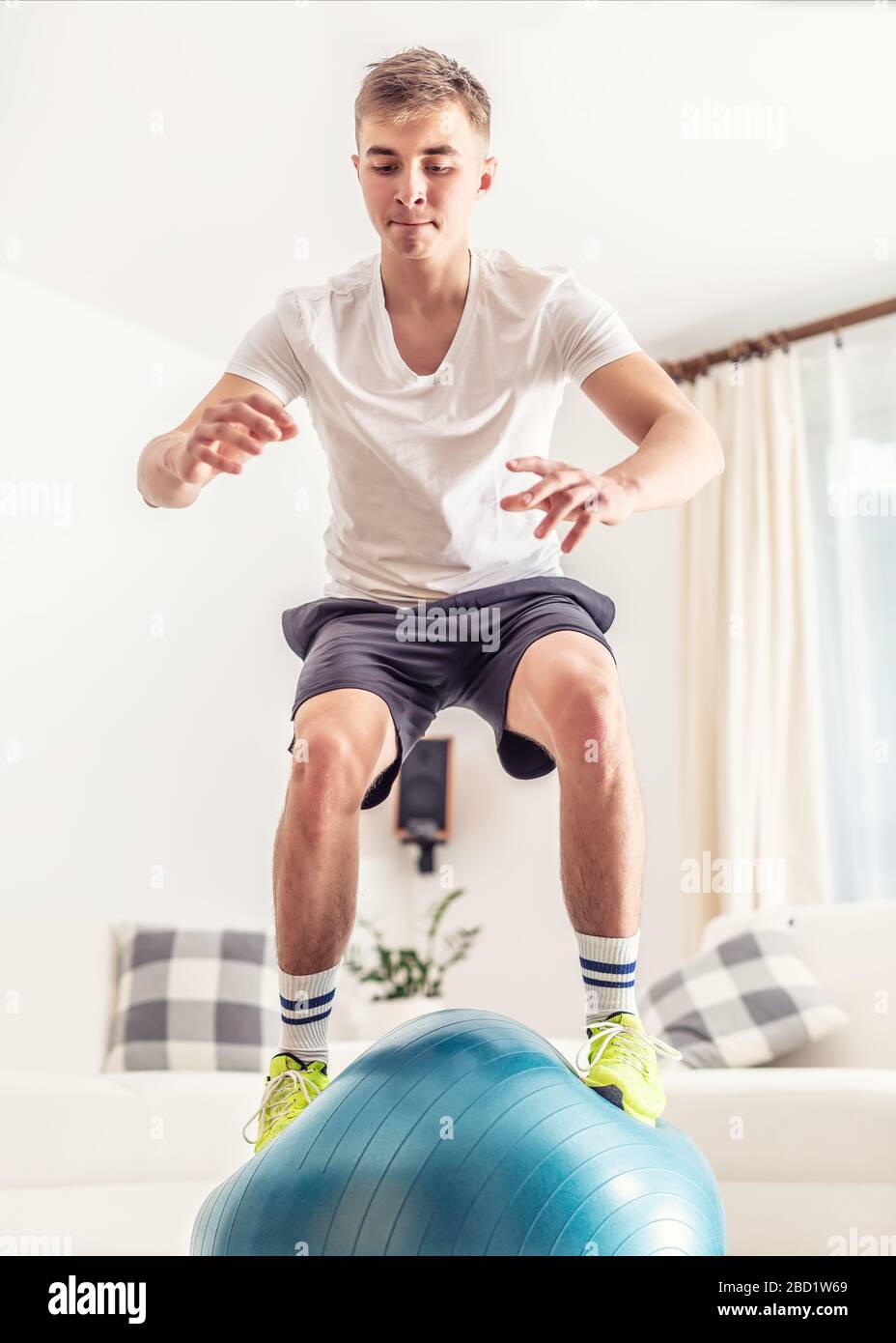 Sportliches, junges Männchen, das beim Heimtraining einen Sprung auf einen blauen Fit Ball beendete, trug T-Shirt, Shorts und Sneaker im skandinavischen Stil auseinander Stockfoto