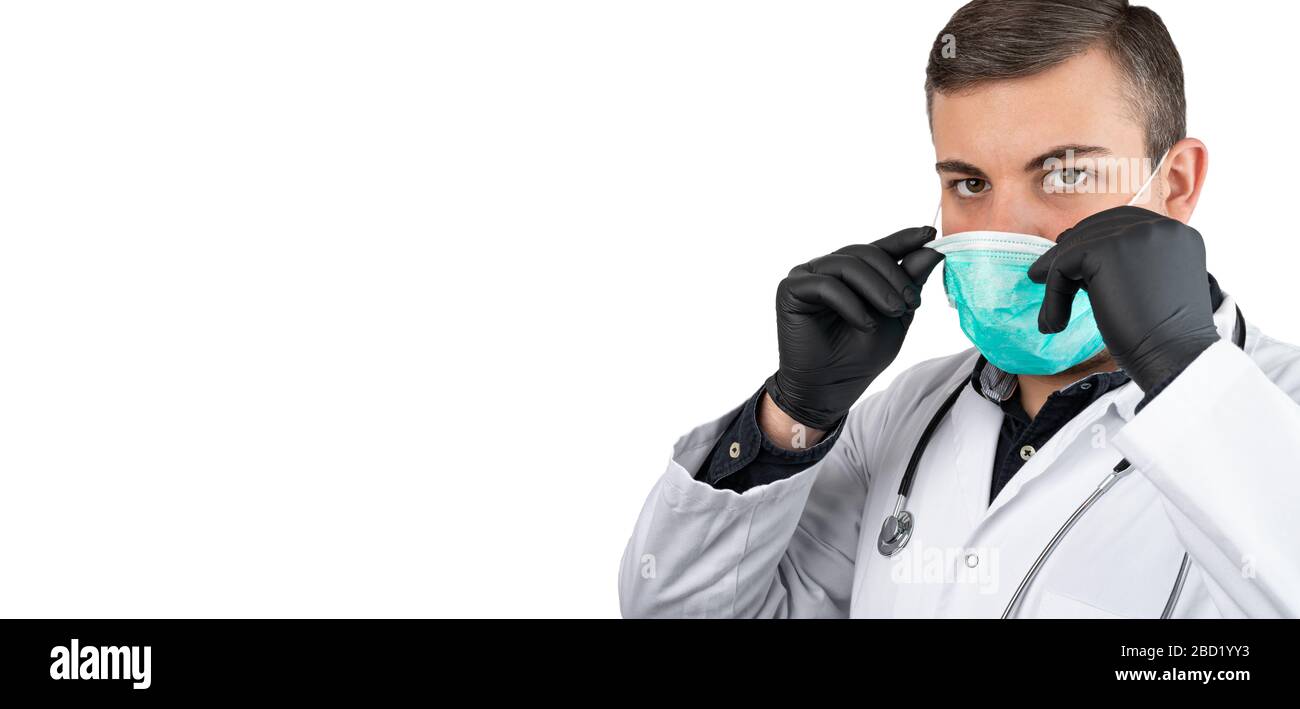 Kaukasischer Mann posiert als Arzt in Weiß insgesamt oder schrubbt mit Stethoskop um einen Hals und schwarze Handschuhe. Aufsetzen einer Gesichtsschutzmaske. Stockfoto