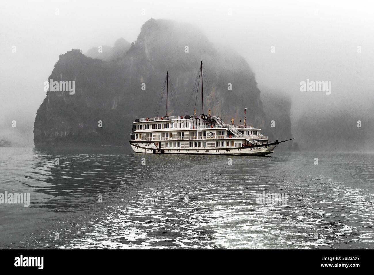 Schwarz-Weiß-Fotografie eines Touristenbootes, das an einem nebligen Tag an einer der vielen Kalksteininseln in der Halong Bay vorbeifährt - Vietnam, Asien Stockfoto
