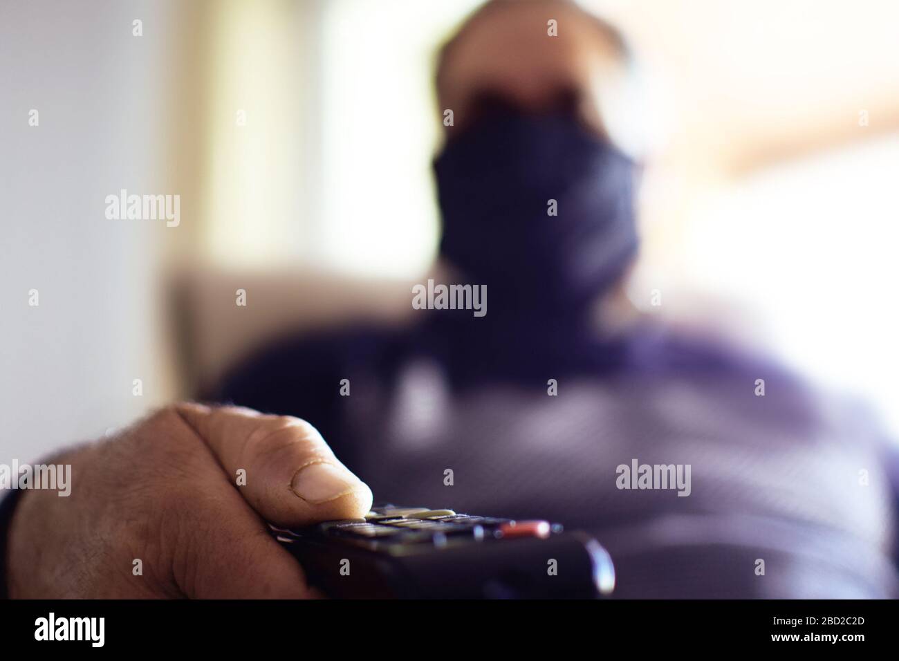 Mann, der während der Quarantäne eine Schutzmaske trägt - Covid 19. Verwenden Sie die Fernbedienung, um das Fernsehprogramm zu ändern. Isolierung zu Hause. Stockfoto