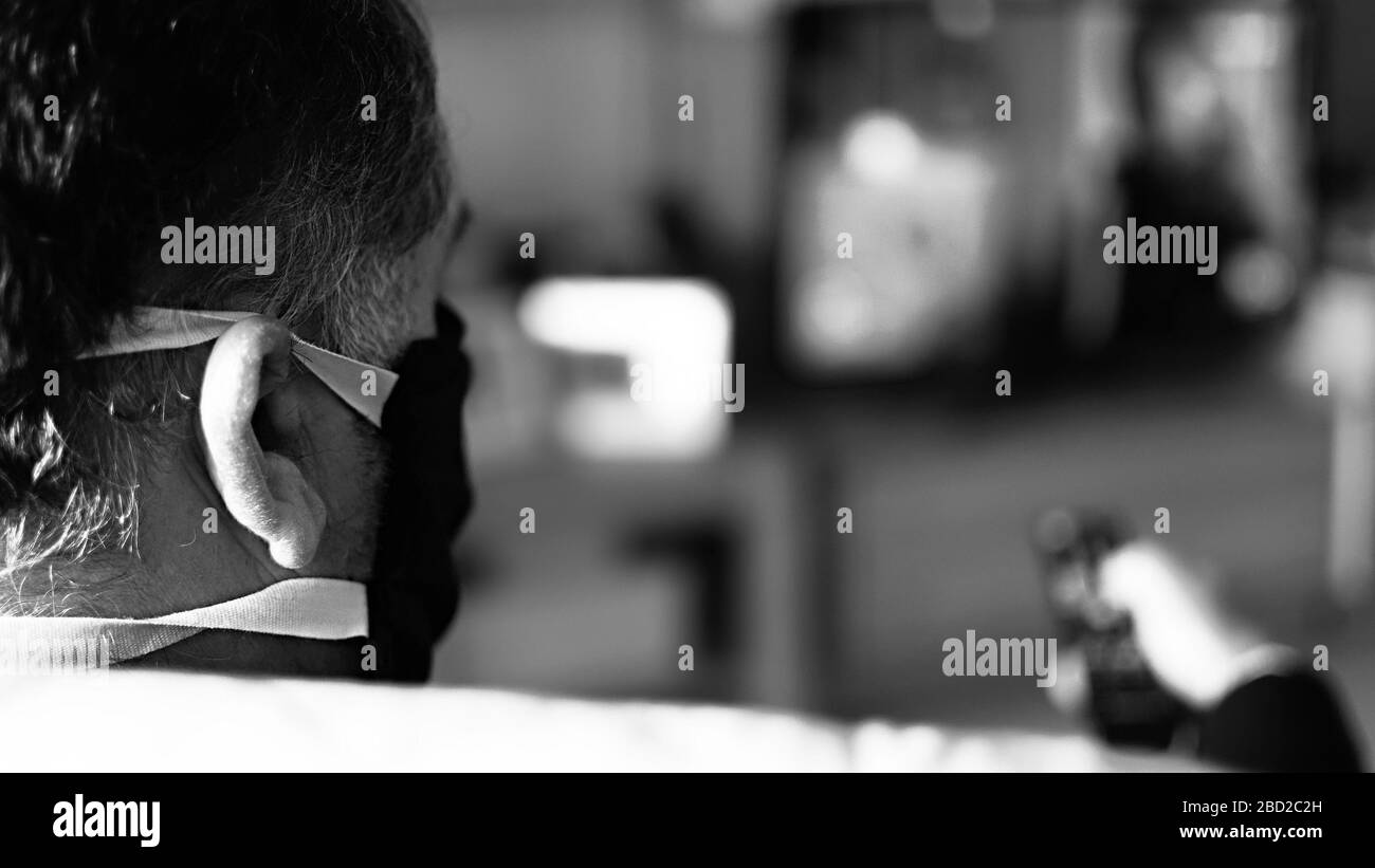 Mann, der während der Quarantäne eine Schutzmaske trägt - Covid 19. Verwenden Sie die Fernbedienung, um das Fernsehprogramm zu ändern. Isolierung zu Hause. Stockfoto