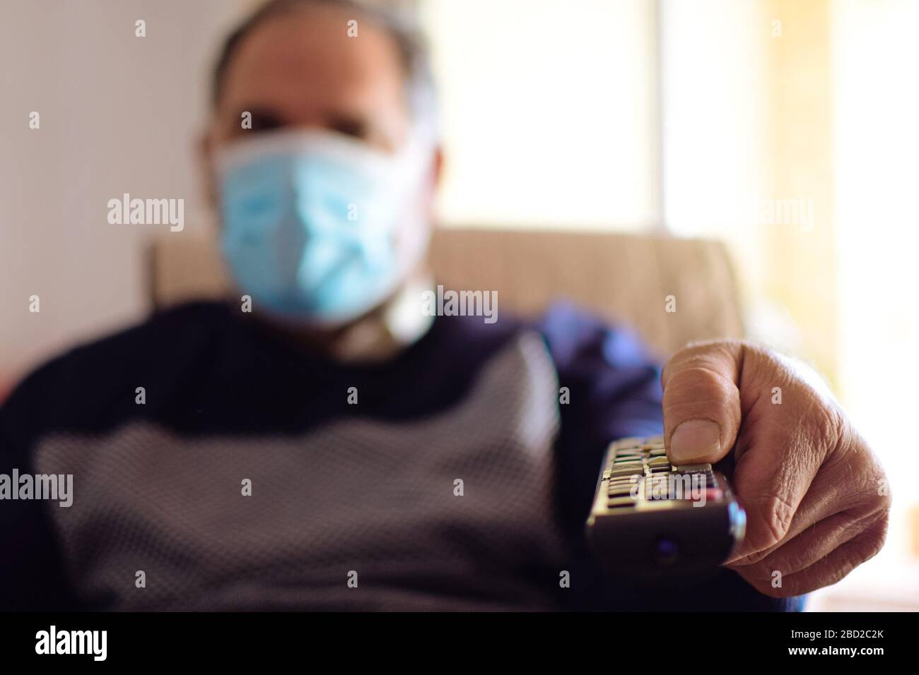 Mann, der während der Quarantäne eine Schutzmaske trägt - Covid 19. Verwenden Sie die Fernbedienung, um das Fernsehprogramm zu ändern. Isolierung zu Hause. Stockfoto