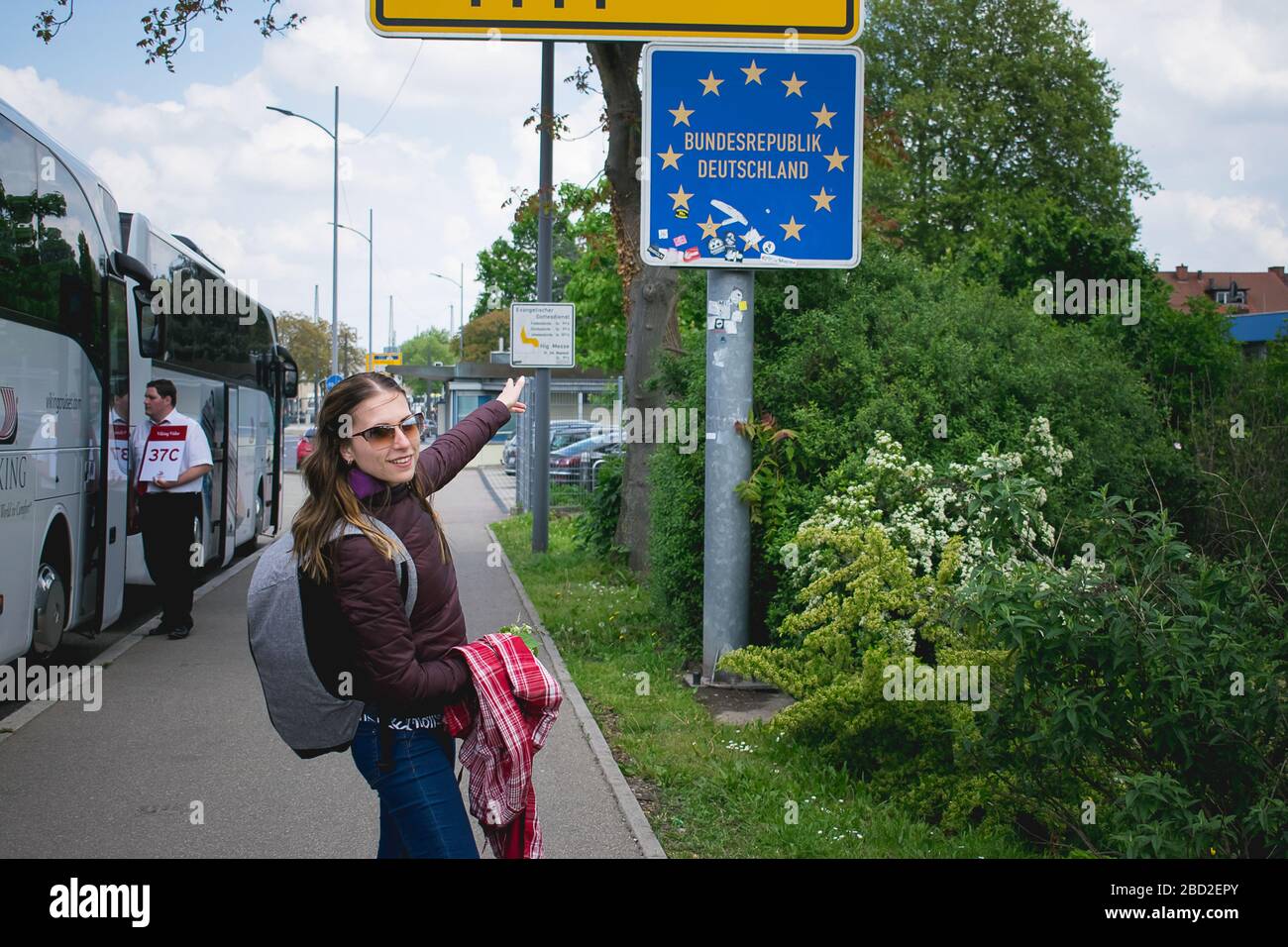 Eine junge Frau überquert eine deutsche Grenze und zeigt auf das Straßenschild. Stockfoto