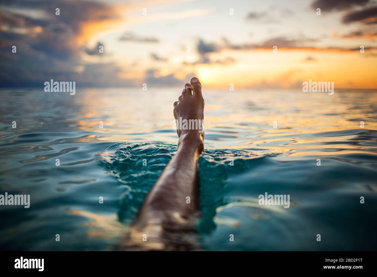 Junge Frau schwebt ihr Bein über dem Wasser, während die Sonne über dem tropischen Ozean untergeht. Stockfoto