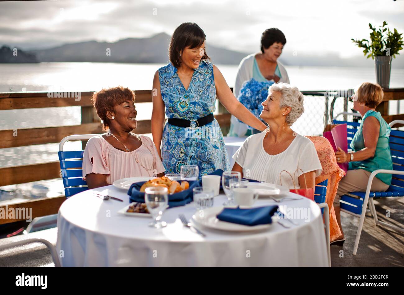 Zwei reife weibliche Freunde, die zum Mittagessen in einem Restaurant am Meer unterwegs sind, lachen mit einem bekannten, der Hallo gesagt hat. Stockfoto