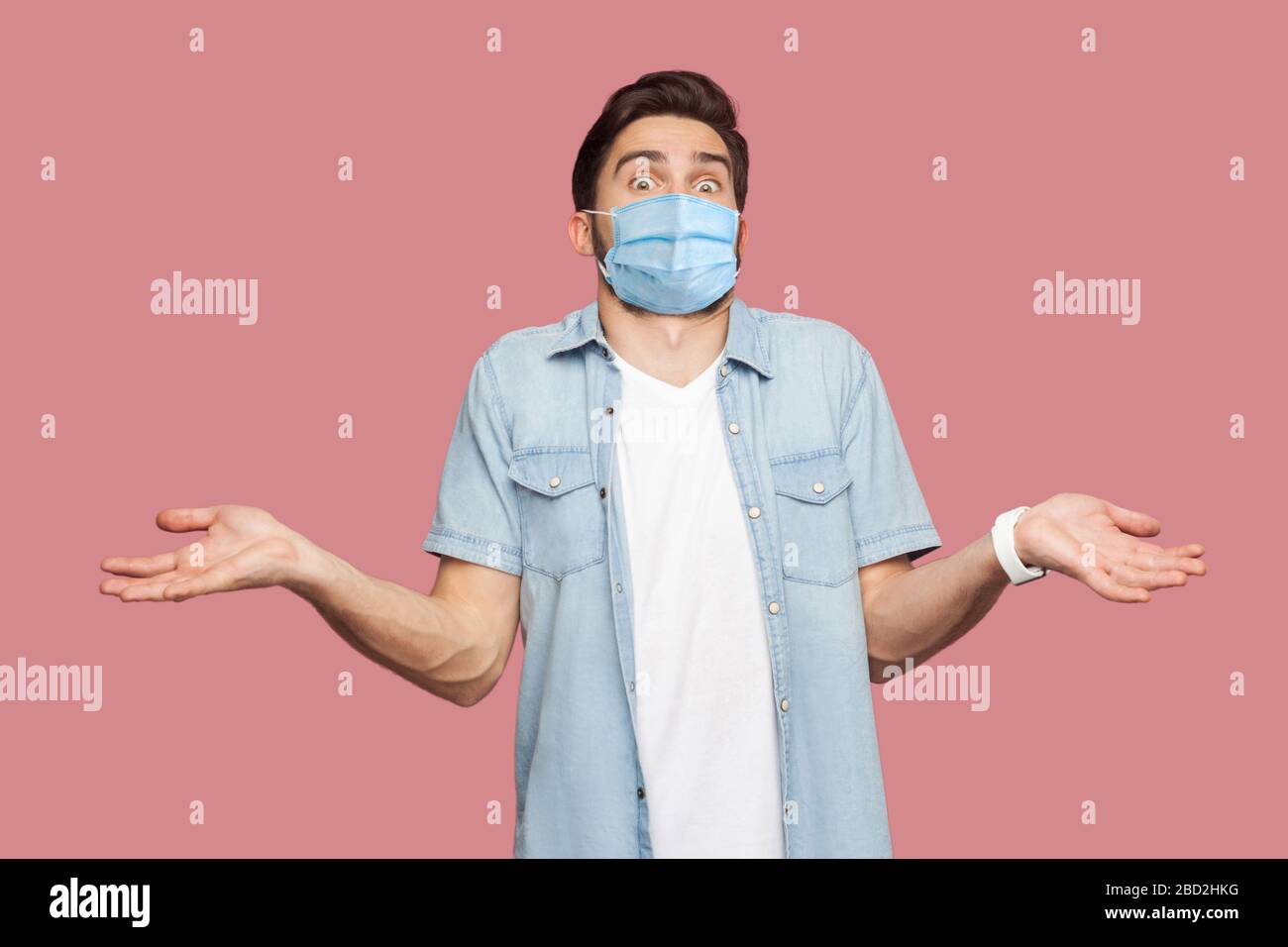 Ich weiß es nicht. Porträt des verwirrten Mannes mit chirurgischer medizinischer Maske in blauem Casual Style Hemd, das mit erhobenen Armen steht und Kamera mit Antwort betrachtet. Stockfoto