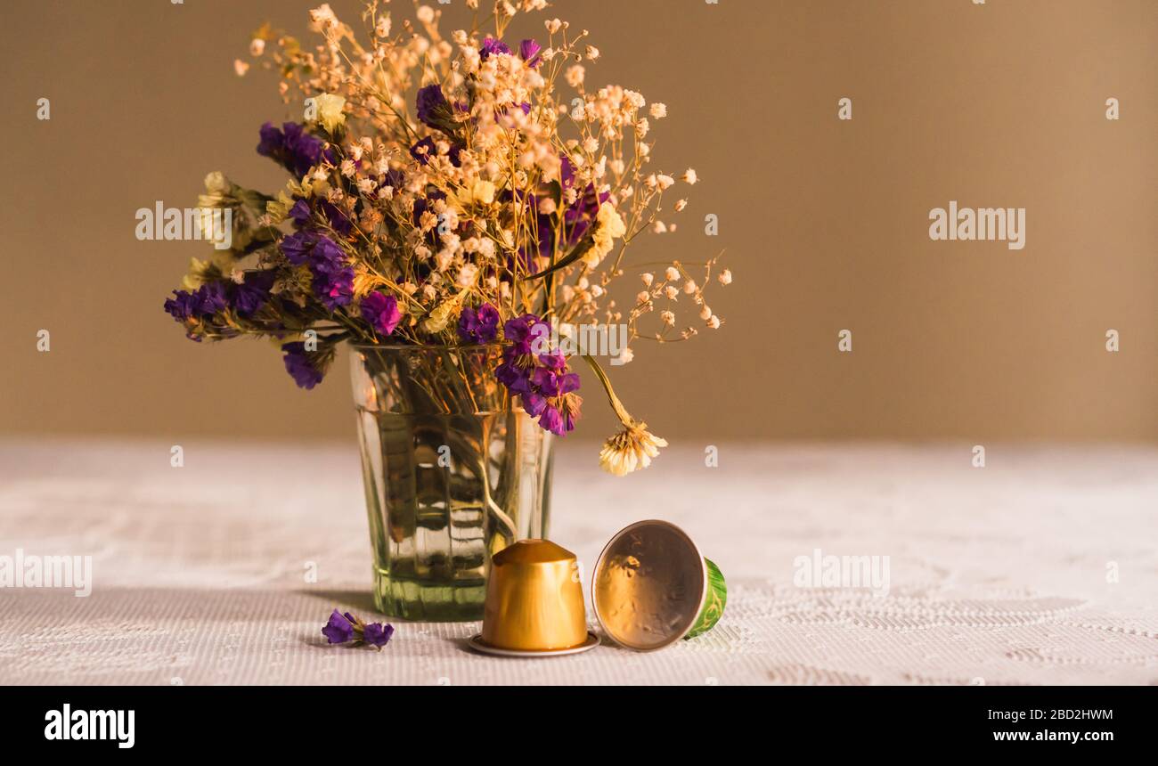 Zwei Nespresso Kaffeekapseln auf dem Tisch vor Die lila und weißen Blüten in einer Glasvase auf Ein weißes Textil auf dunklem Hintergrund Stockfoto