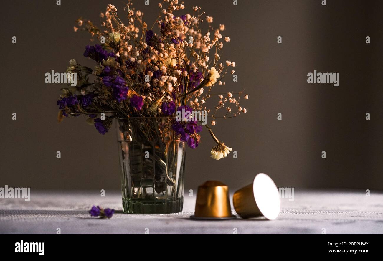 Zwei Nespresso Kaffeekapseln auf dem Tisch vor Die lila und weißen Blüten in einer Glasvase auf Ein weißes Textil auf dunklem Hintergrund Stockfoto