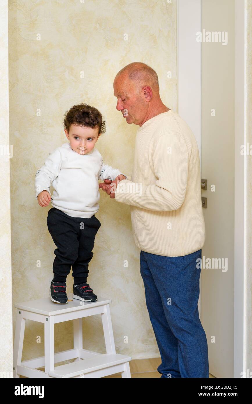 Lustiger kleiner Junge springt von Stepladder und hält die Hände mit Großvater. Glückliche Familie zu Hause, die einfache Momente genießt. Stockfoto