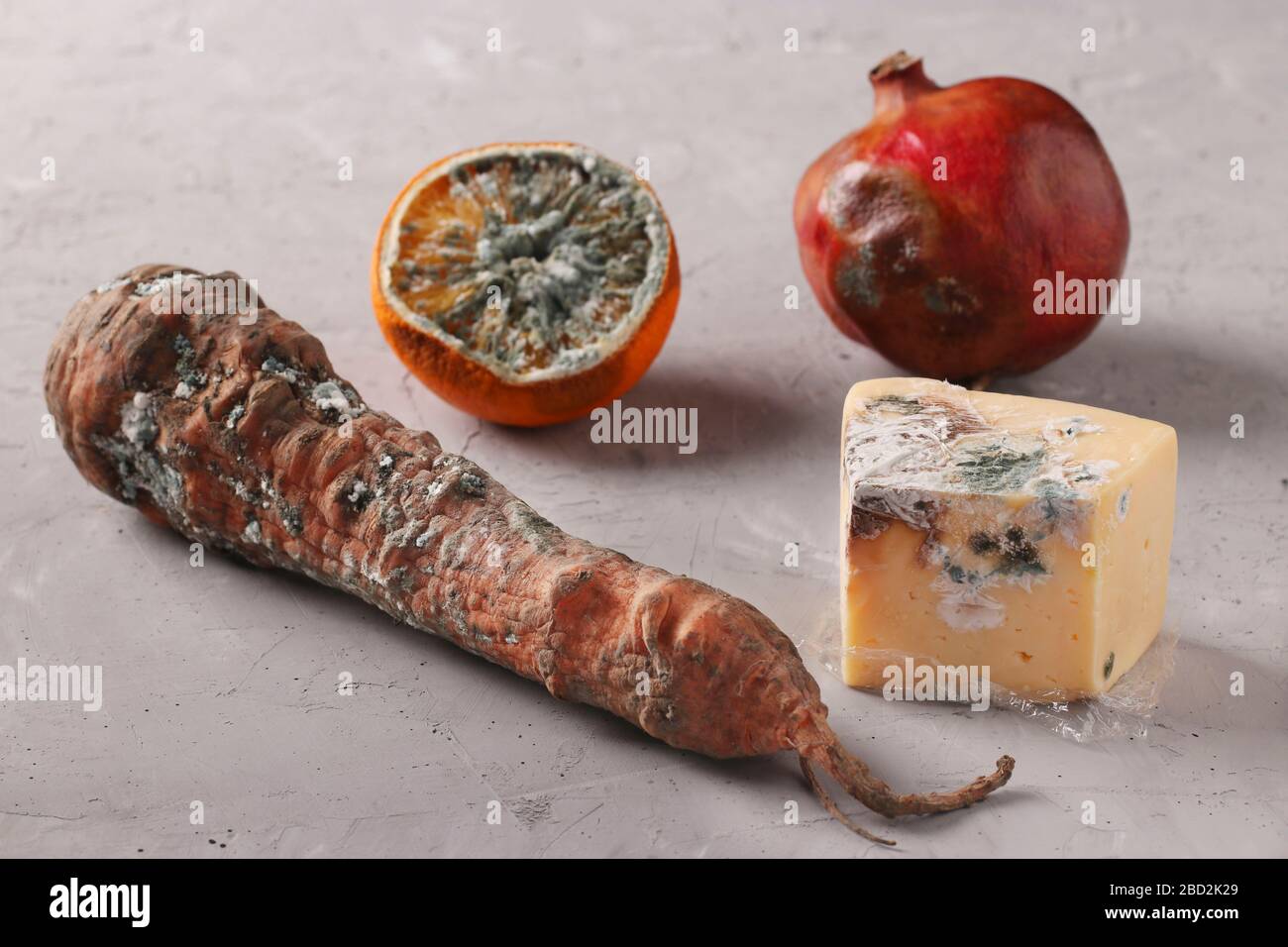 Verdorbene faule Speisen mit Schimmel: Halbe Orange, Granatapfel, Karotten und Hartkäse auf grauem Grund Stockfoto