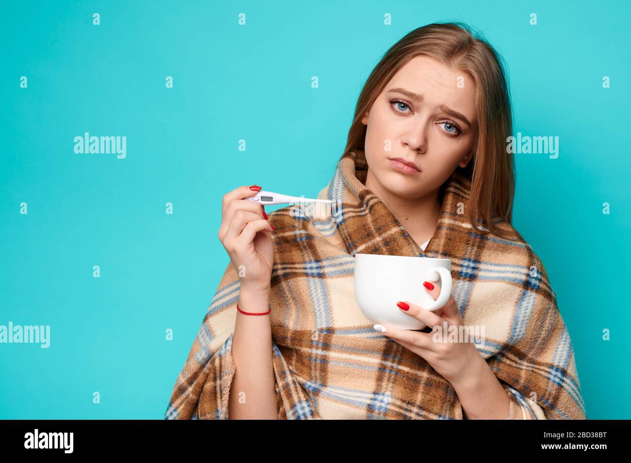 Eine Frau in einem Pullover und eine Tasse in den Händen misst die Temperatur mit einem Thermometer. Virus, Grippe, krank, Kovid19, Coronavirus Konzept Stockfoto