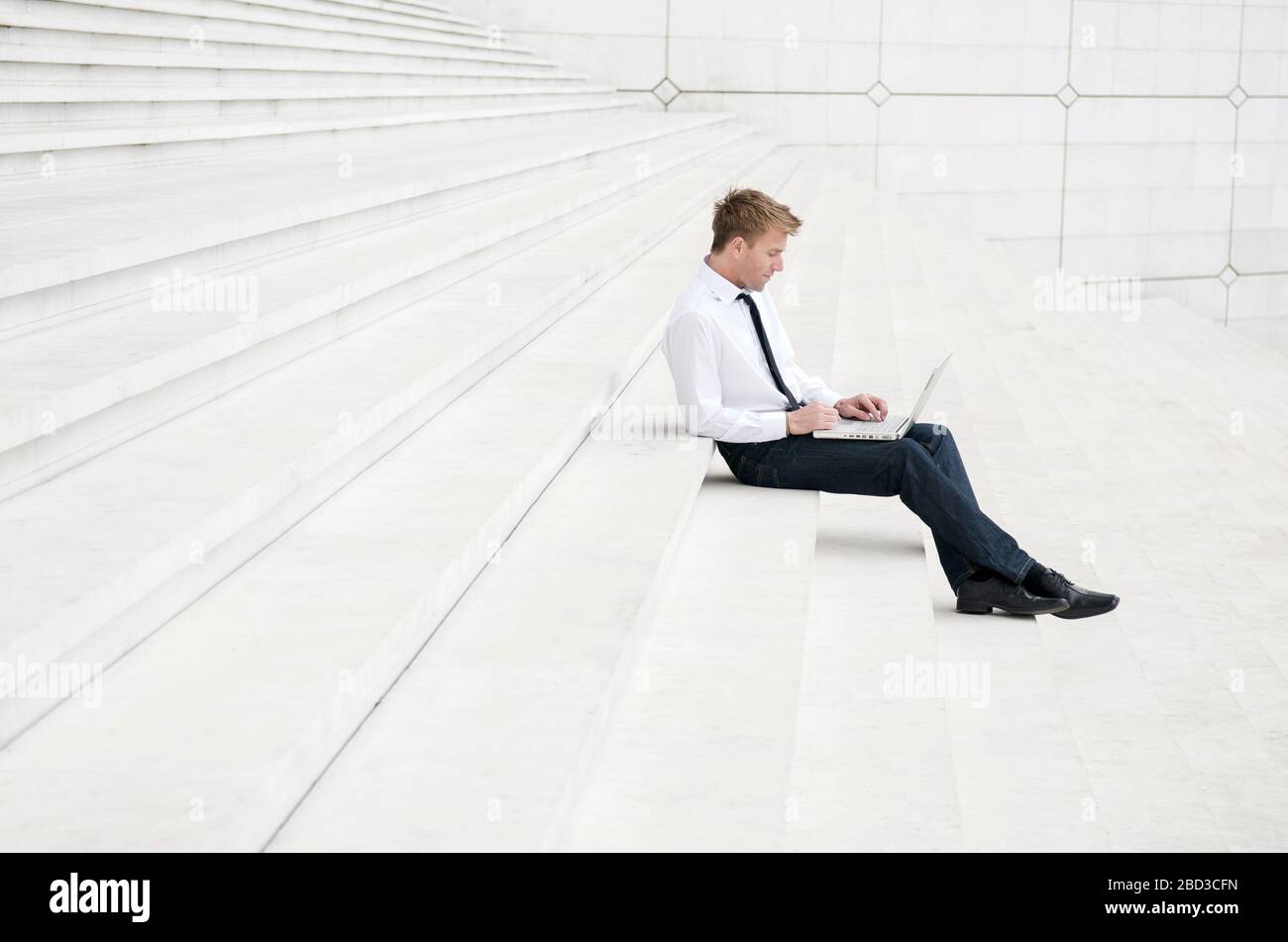 Junger Mann in einem eleganten lässigen weißen Hemd und Jeans sitzt im Freien auf minimalistischen Marmorstufen mit seinem Laptop-Computer Stockfoto