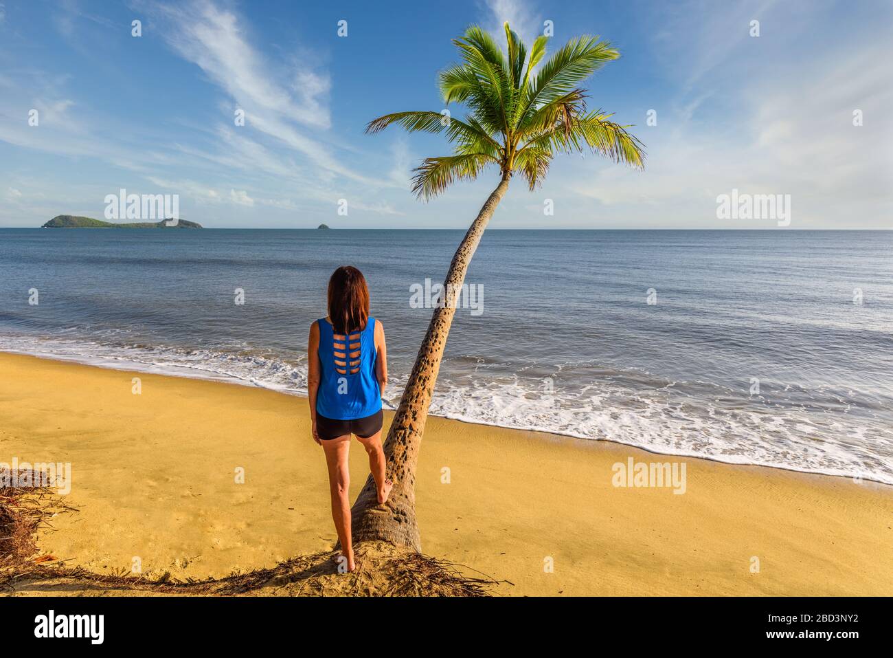 Blick auf einen einzigen weiblichen Tourist, der in der Nähe einer Kokospalme am Clifton Beach mit Blick auf den Pazifischen Ozean und die Doppelinsel in Queensland steht. Stockfoto