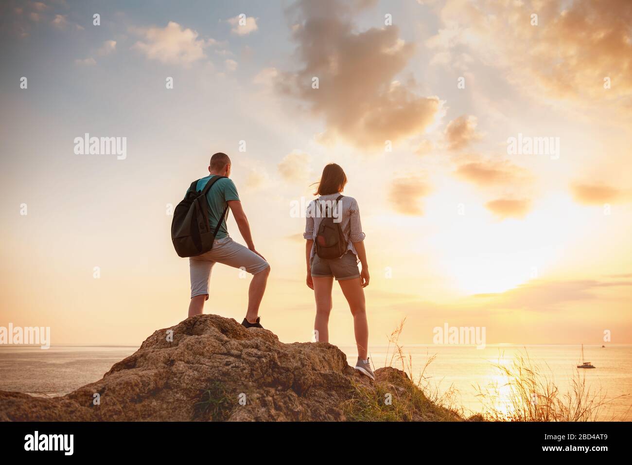 Zwei junge Wanderer, männlich und weiblich, stehen auf einem großen Felsen und blicken auf das Meer und den Himmel bei Sonnenuntergang Stockfoto