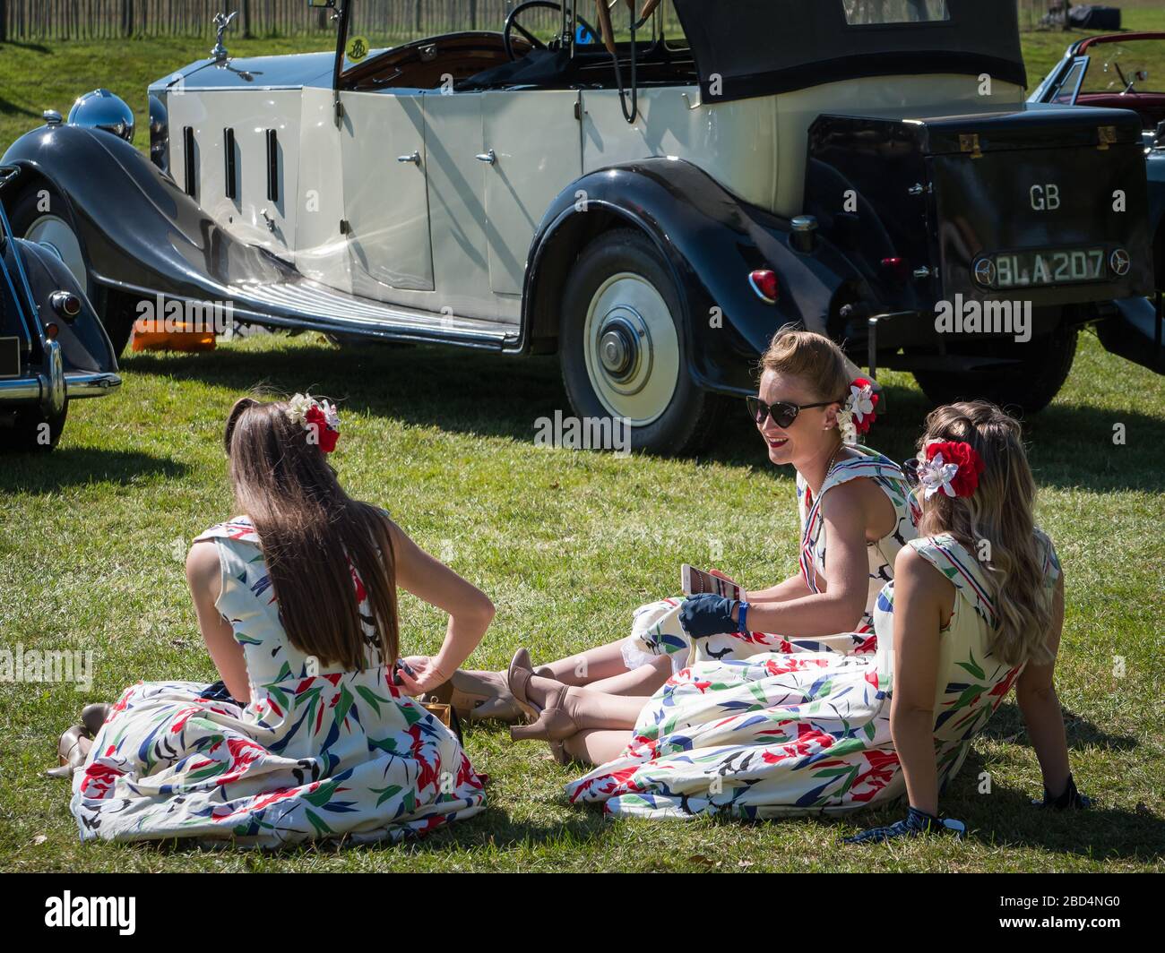 Drei Frauen im Vintage-Kleid sitzen im Sonnenschein, GRRC-Gehäuse auf der Lavant Straight, Goodwood Revival 2019 West Sussex UK Stockfoto