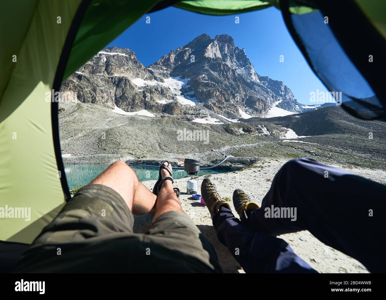 Männliche Beine im Zeltlager mit schneebedeckten Bergen im Hintergrund.  Zwei Reisende, die im Touristenzelt liegen und die Aussicht auf die schönen  felsigen Hügel der Alpen genießen. Konzept für Reisen, Wandern und Campen