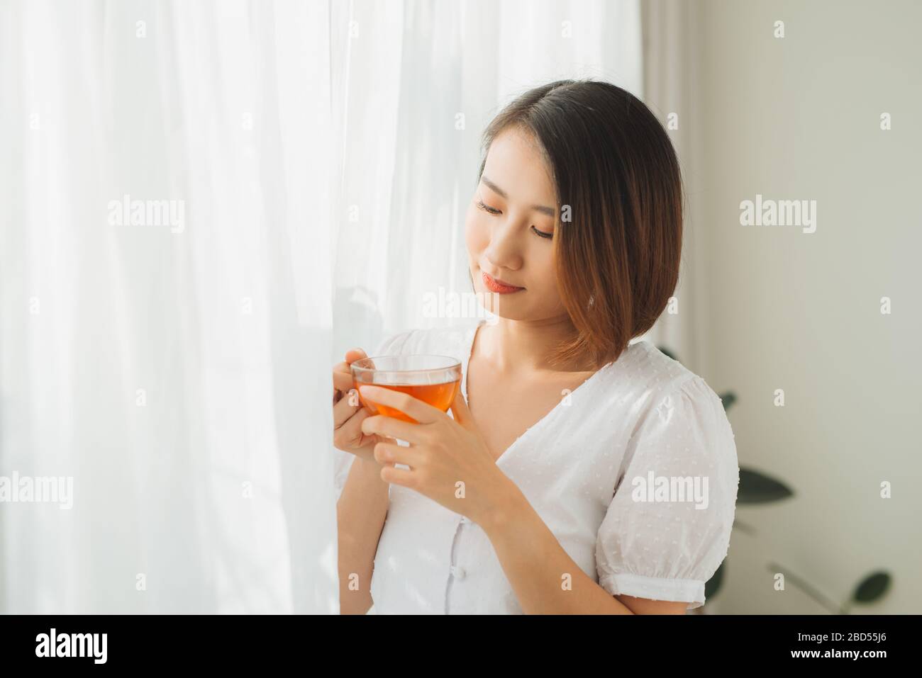 Schöne Frau, die am Fenster steht und ein Glas heißen Tee in der Hand hat. Konzept für Nachmittagstee. Stockfoto