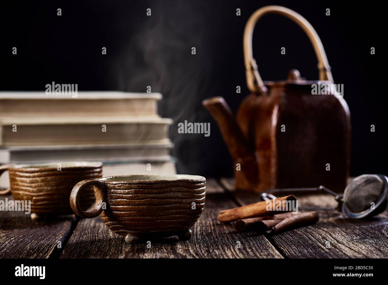 Nahaufnahme des Stilllebens mit Keramikbecher aus Tee oder Kaffee, Teekanne und Zimt. Alte Holztischplatte, Rauch vor Büchern und verschwommener schwarzer Bac Stockfoto