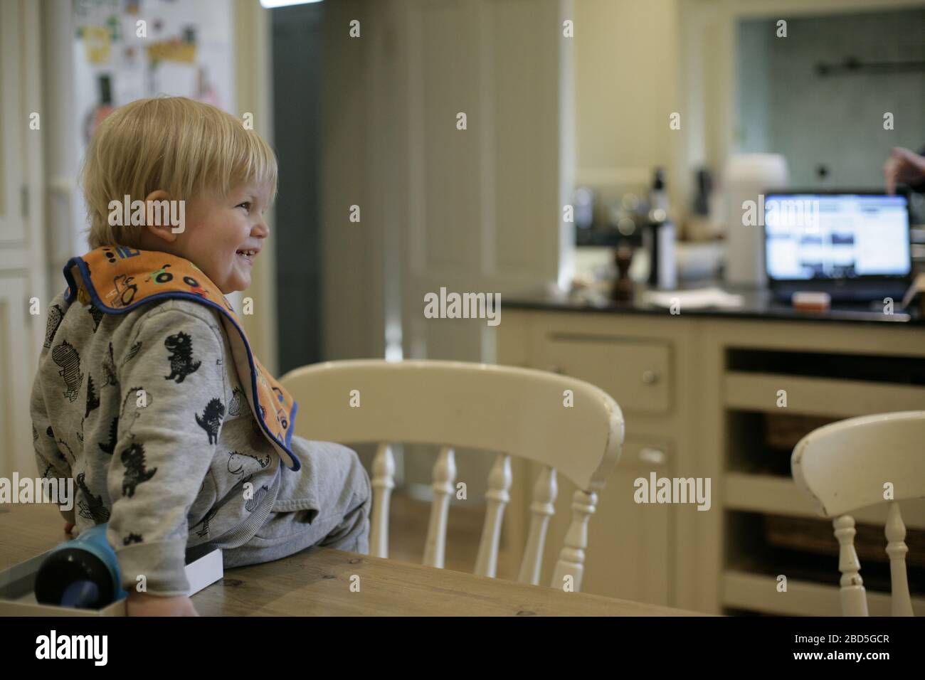Das Kind sitzt zu Hause auf dem Tisch, während die Mutter während der Zeit der Selbstisolierung während der COVID-19-Pandemie 2020 in der Küche arbeitet Stockfoto