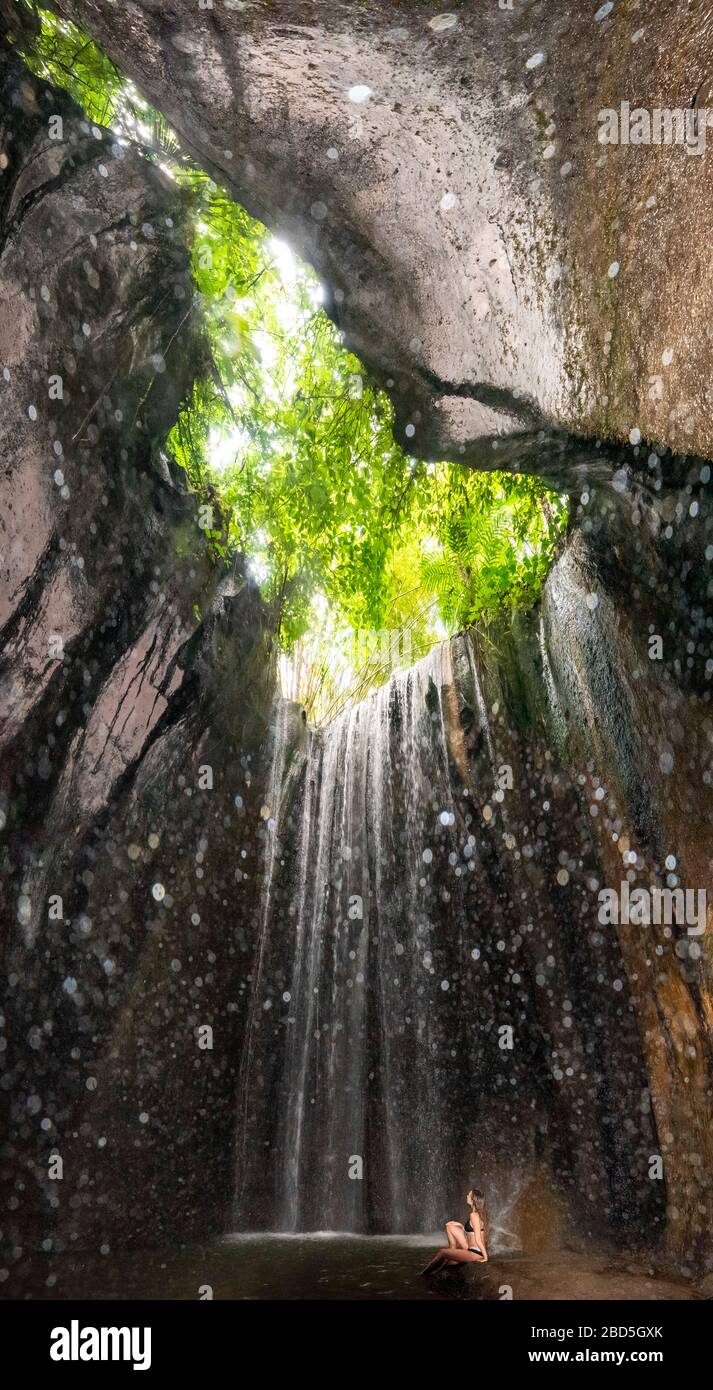 Vertikales Porträt einer Frau, die unter den Tukad Cepung-Wasserfällen in Bali, Indonesien sitzt. Stockfoto
