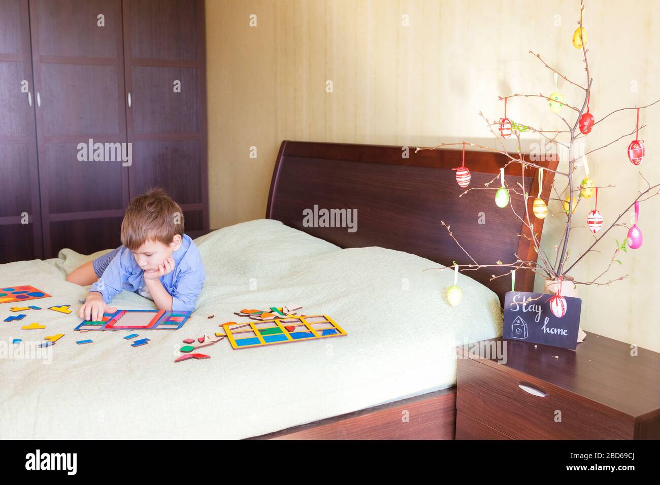 Langweiliger Junge sammelt Holz Puzzle sitzen auf dem Bett zu Hause Innenraum. Osterdekoration im Hintergrund und Text zu Hause bleiben. Zu Ostern zu Hause sein Konzept Stockfoto