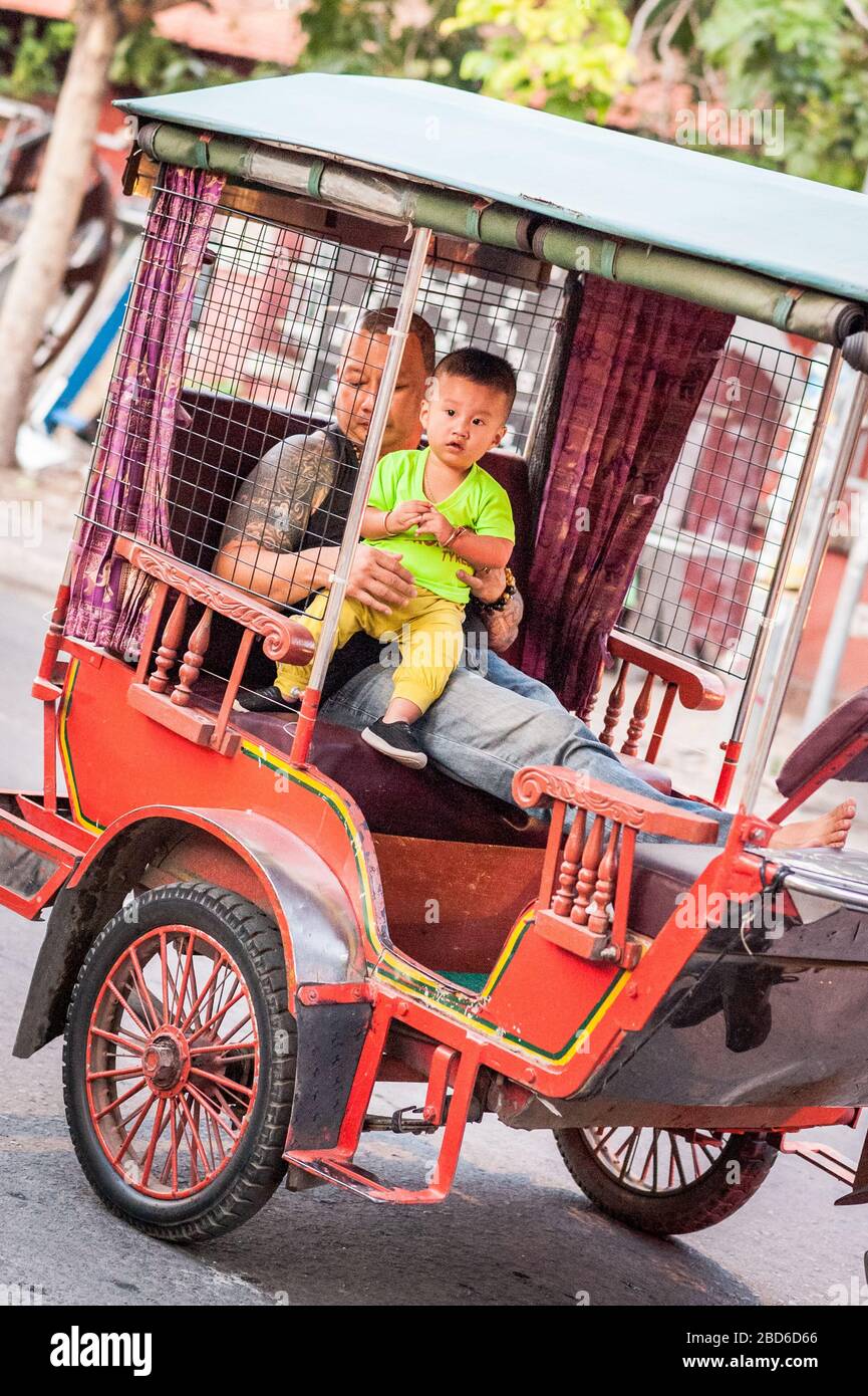 Ein Vater hält sein Baby fest, während sie sich im Rücken einer traditionellen Rikscha setzen und sich durch die belebten Straßen von Phnom Penh machen. Stockfoto