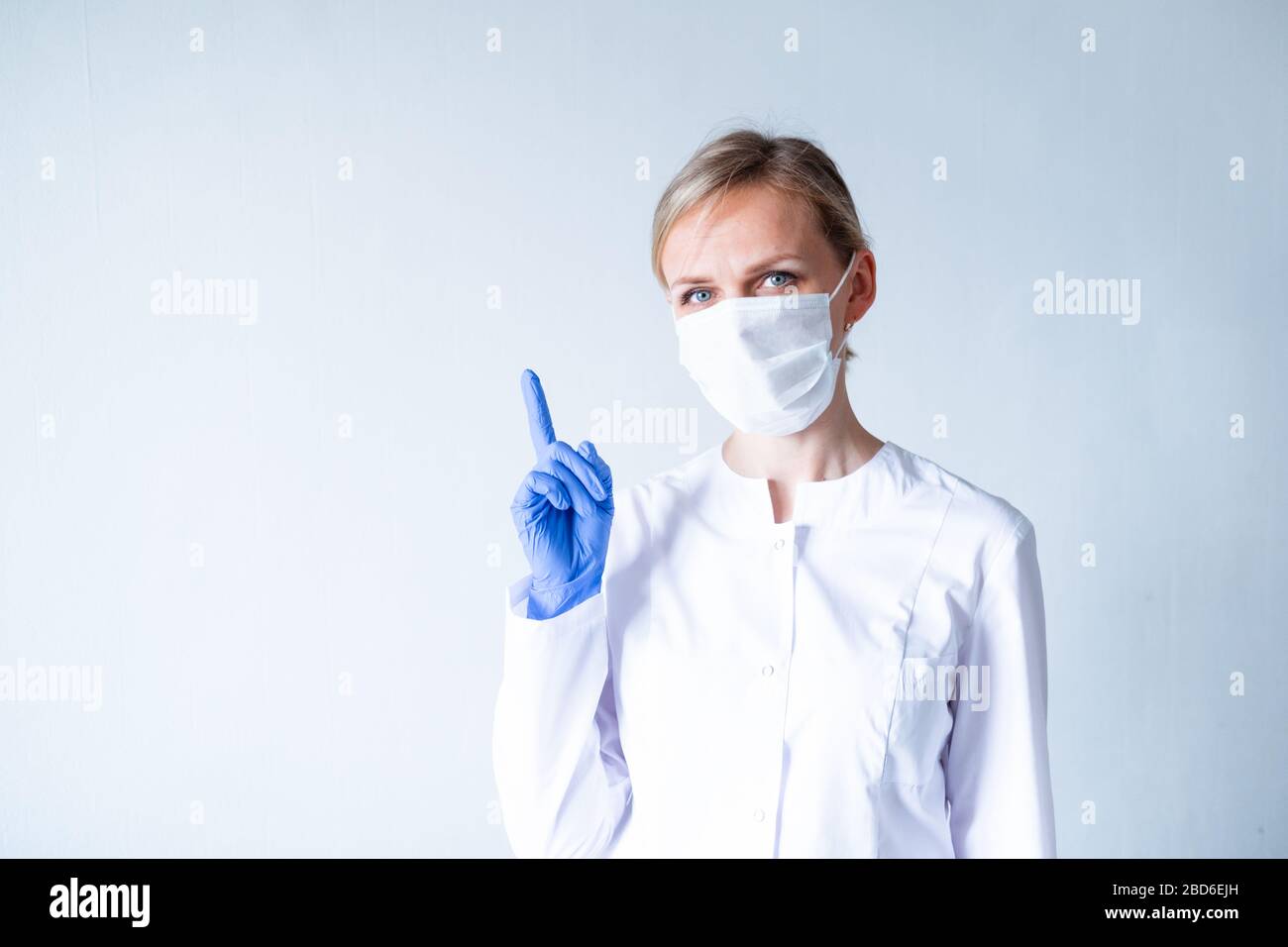 Achtung. Blonde Ärztin in weißer Uniform, blaue Schutzhandschuhe und medizinische Maske, die den Finger über Grau zeigt. Stockfoto