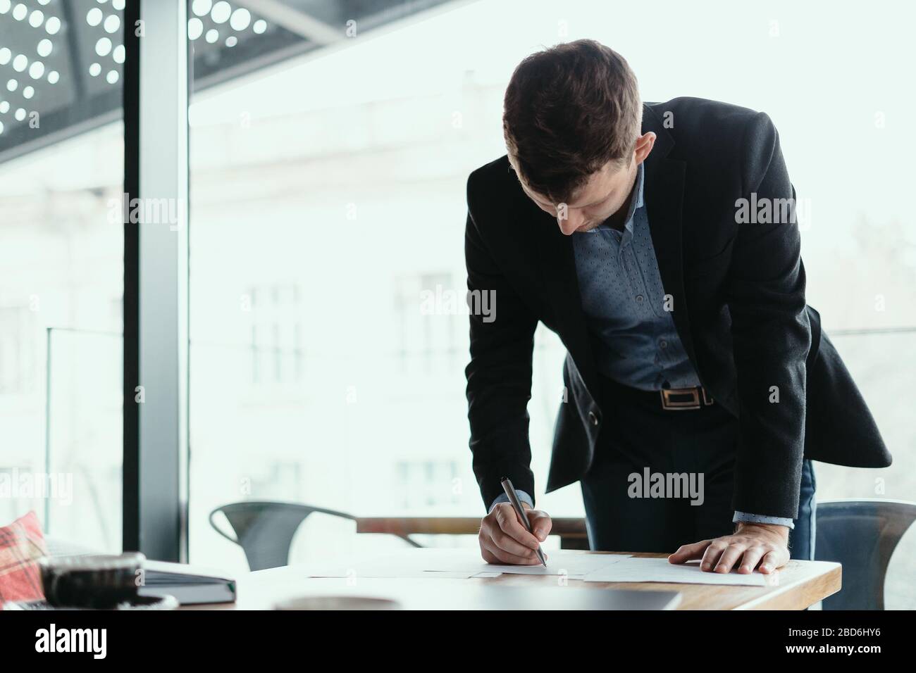 Selbstbewusster junger Business man unterschreibt Dokumente in einem modernen Büro mit Fenster im Hintergrund. Stift in der Hand, Papier auf dem Holztisch Stockfoto