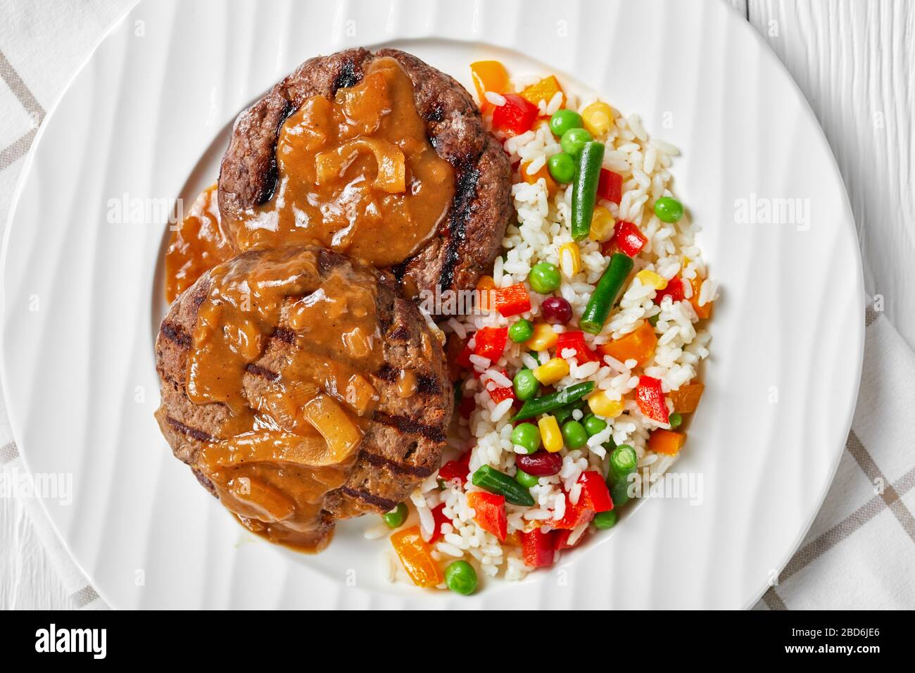 Gegrillte Hamburger Steaks mit Zwiebelgravy und Reis gemischt mit Gemüse auf einem weißen Teller auf einem Holztisch, flach gelegt, Nahaufnahme Stockfoto