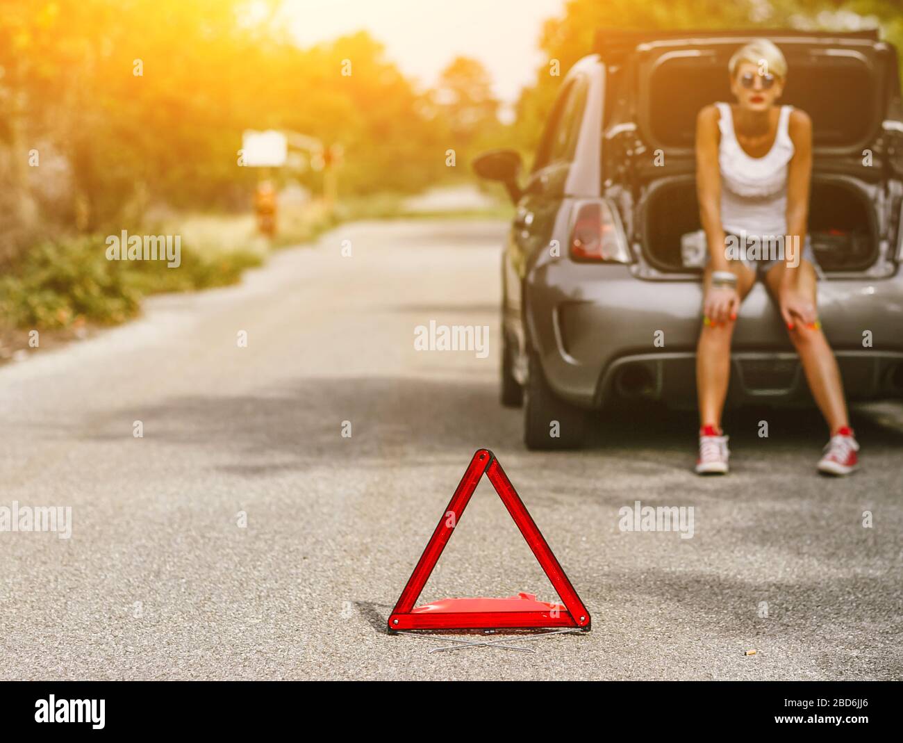 Junge schöne Frau spection einen Hinterreifen auf der Seite der Straße. Auto mit Problemen und roten Dreieck, um andere Verkehrsteilnehmer zu warnen. Stockfoto