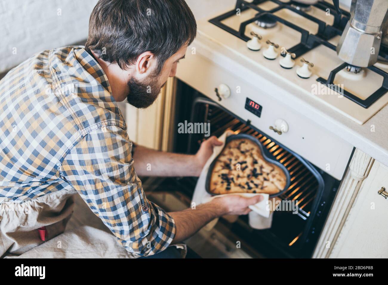 Bärtiger junger Mann, der herzförmige Beerenkuchen aus dem Ofen nimmt. Zu Hause kochen und lächeln. Stockfoto