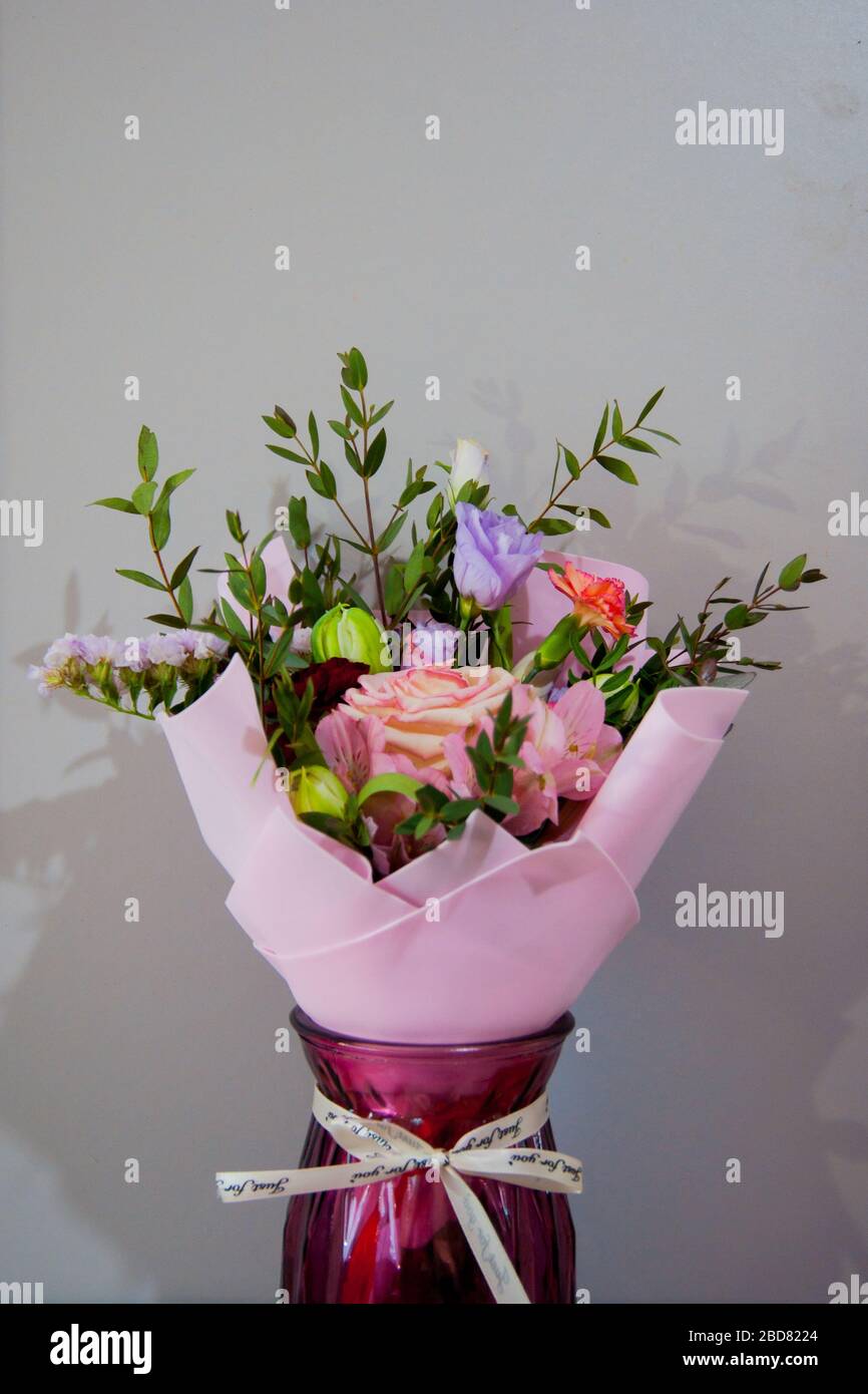 Ein zartes Blumenstrauß verschiedener Blumen in einem hellrosa Wrapper steht in einer weinroten Vase auf grauem Grund. Vertikales Foto. Stockfoto
