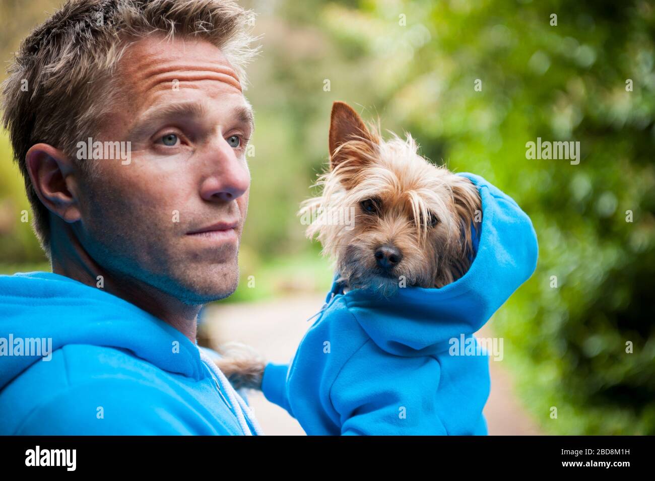 Hundebesitzer trägt seinen schroffigen kleinen besten Freund in passenden blauen Hoody Sweatshirts im Freien vor dem Grün stehen Stockfoto
