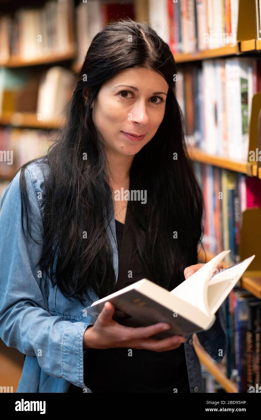 Porträt einer jungen Frau, die in der Bibliothek ein offenes Buch hält und dabei vor die Kamera lächelt Stockfoto